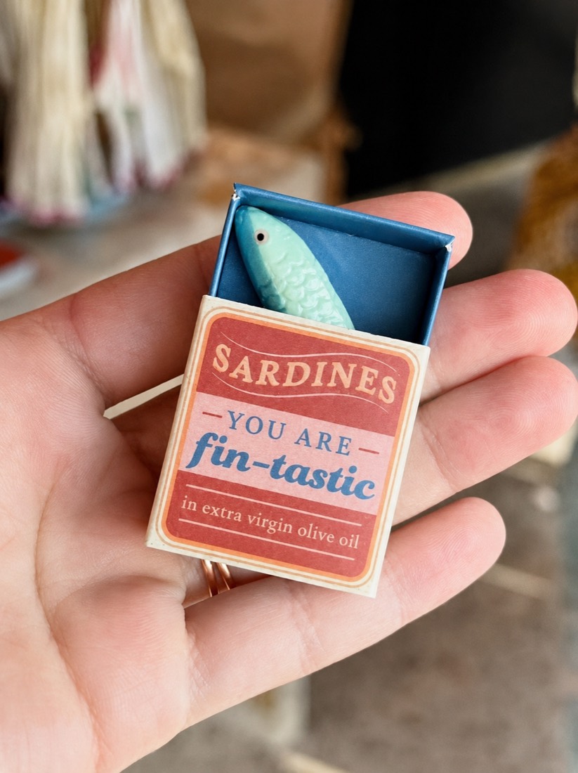 A hand holding a small sardine-tin matchbox with a ceramic fish tucked inside
